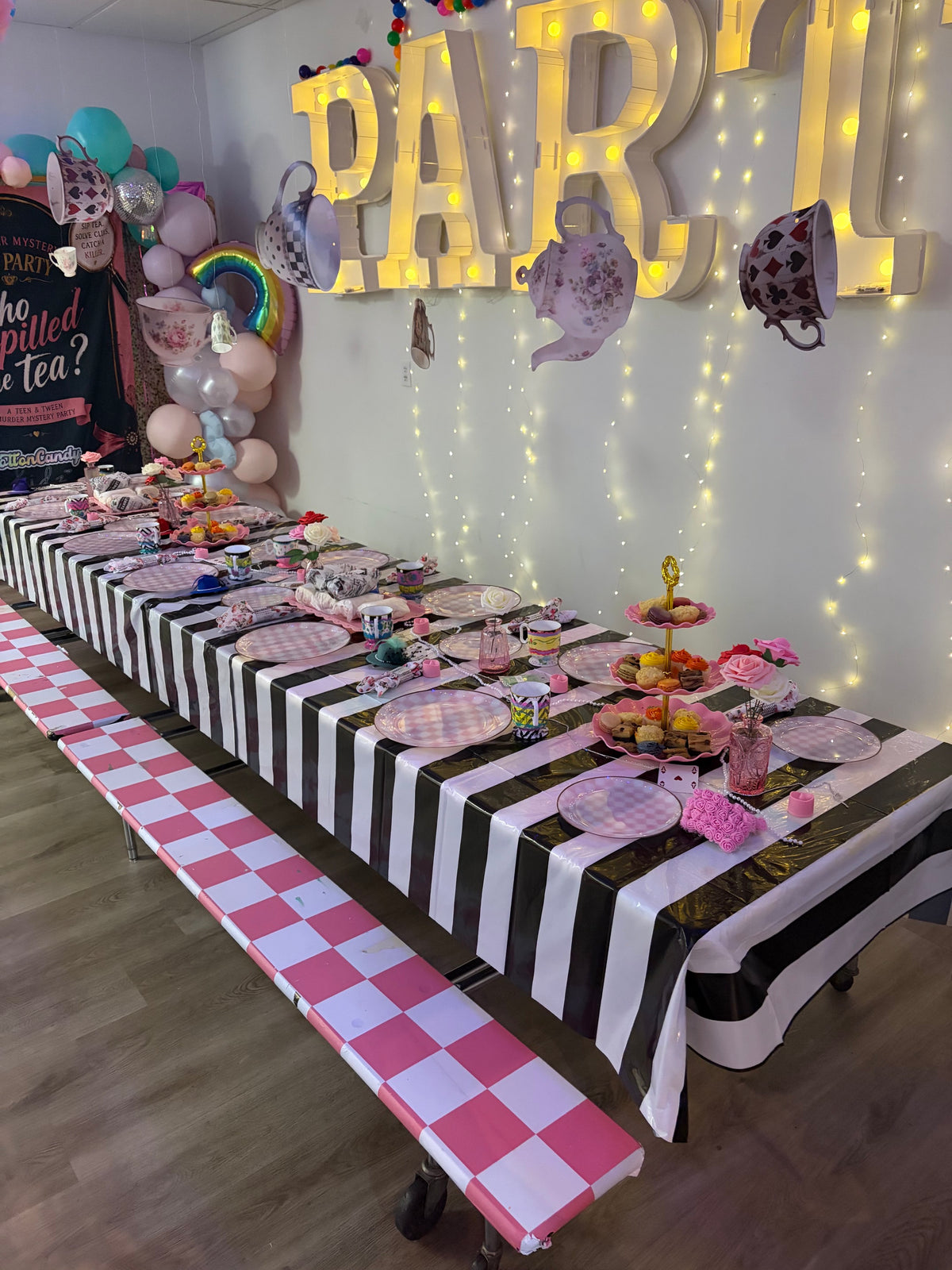 A long party table decorated with a black and white striped tablecloth, pink and white checkered runner, matching plates, teacups, and tea-time treats, set against a wall with large illuminated 'PARTY' letters and hanging teapot decorations.