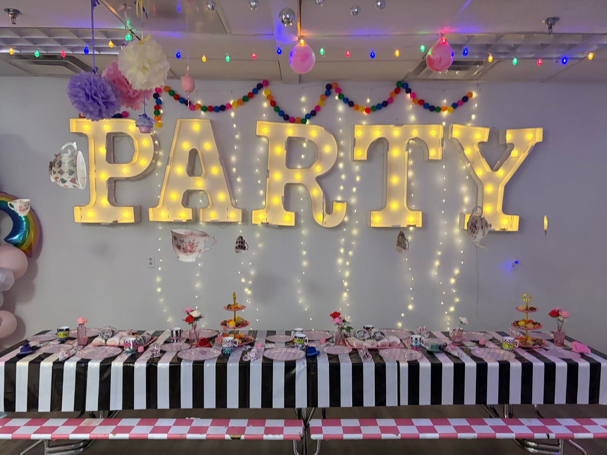 A decorated party setup with large illuminated marquee letters spelling PARTY on the wall, colorful string lights, pom-poms, balloons, and a long table covered in a black-and-white striped tablecloth with party accessories.